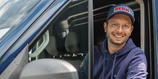 Ein lächelnder Mann sitzt am Fenster eines blauen Fahrzeugs und trägt eine Kappe mit dem Logo einer Heizungsfirma.