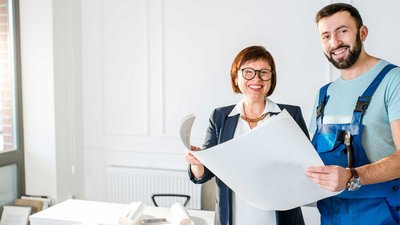 Ein Mann in Arbeitskleidung steht neben einer Frau, die eine Zeichnung in der Hand haelt