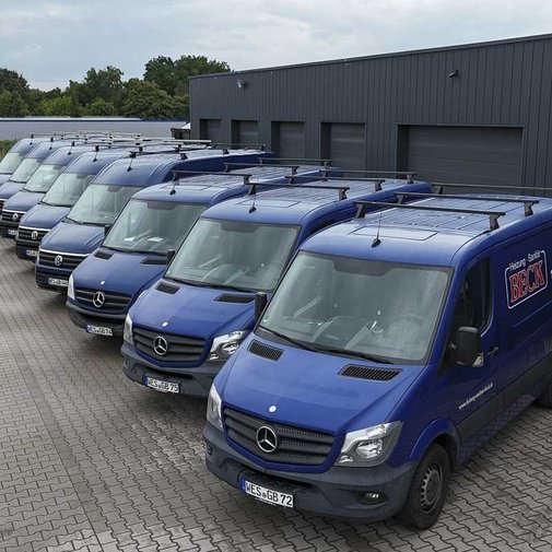 Eine Reihe von blauen Lieferwagen steht ordentlich auf einem gepflasterten Parkplatz vor einem modernen Gebäude.
