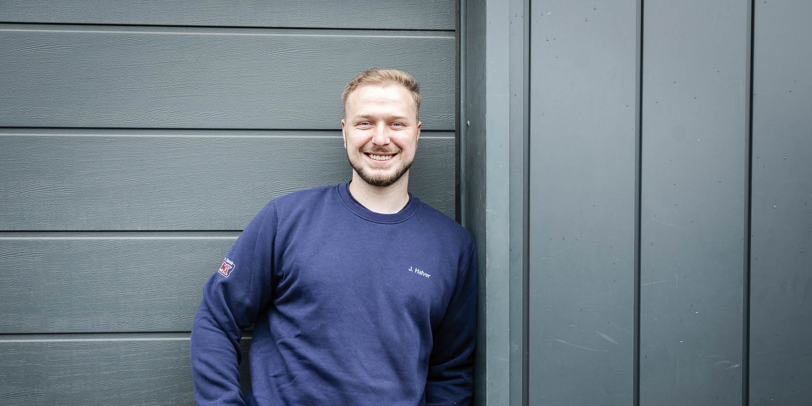 Der junge Mann lächelt fröhlich, während er an einer schlichten, grauen Wand lehnt, die ihm einen modernen Look verleiht.