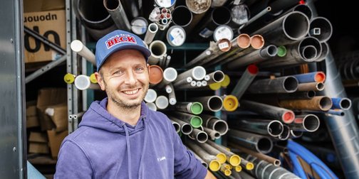 Ein lächelnder Mann in einem blauen Kapuzenpullover steht vor einem Regal voller bunter Rohre und Materialien.