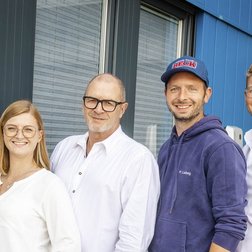 Vier Personen stehen lächelnd vor einem modernen Gebäude mit blauer Fassade und großen Fenstern.