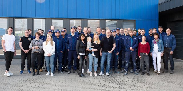 Eine Gruppe von Mitarbeitern steht vor einem blauen Gebäude, lächelnd und in Arbeitskleidung, während einige Familienmitglieder dabei sind.