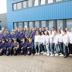 Eine große Gruppe von Mitarbeitern steht vor einem blauen Gebäude, gekleidet in dunkle und helle Kleidung, mit einem freundlichen Lächeln.