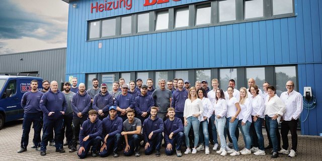 Eine Gruppe von Mitarbeitern steht vor einem blauen Gebäude, lächelnd und in Arbeitskleidung, bereit für ein Gruppenfoto.