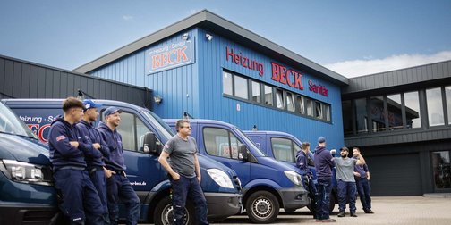 Eine Gruppe von Handwerkern steht entspannt vor einem blauen Gebäude mit dem Schriftzug 'Heizung Beck'.